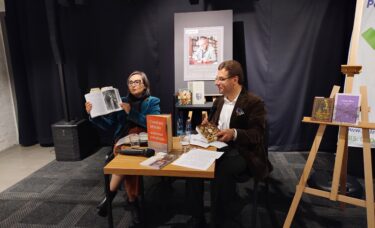 Agnieszka Kosińska unosi książkę, siedzi przy stoliku z Nikitą Kuznetsovem. W tle plakat ze spotkania i książki o Miłoszu