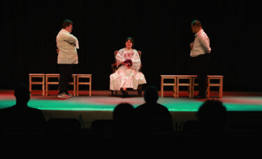 trójka aktorów na scenie, dwóch kelnerów patrzy na pchłe szachrajkę siedzącą na krzesełku