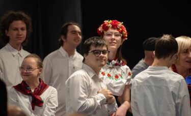 Zbliżenie na grupę wykonawców na scenie. W centrum chłopak w okularach i białej koszuli uśmiecha się, trzymając złożone dłonie na piersi. Obok niego stoi dziewczyna w tradycyjnym stroju ludowym z czerwonym wiankiem na głowie.