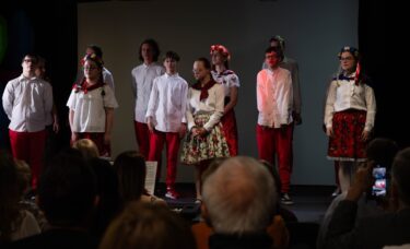 Grupa młodzieży w strojach ludowych i biało-czerwonych ubraniach stoi w rzędzie na zaciemnionej scenie podczas występu przed publicznością.