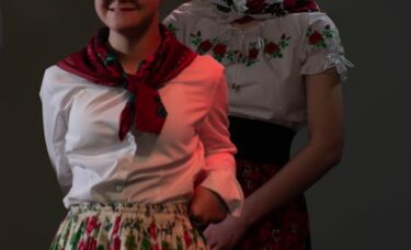Dwie dziewczyny na scenie w tradycyjnych strojach z motywami kwiatowymi. Dziewczyna na pierwszym planie uśmiecha się, ma na sobie okulary i czerwoną chustę. Za nią stoi druga dziewczyna w dużym, czerwonym wianku.