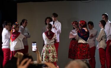 Grupa młodzieży zebrana na scenie w kręgu podczas występu. Chłopak pośrodku trzyma mikrofon i mówi, podczas gdy reszta uczestników w strojach ludowych słucha go z założonymi rękami.