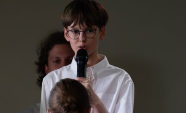 Zbliżenie na chłopaka w okularach i białej koszuli, który śpiewa lub mówi do mikrofonu na scenie. Przed nim tyłem stoi dziewczyna z zaplecionym warkoczem i czerwoną chustą.