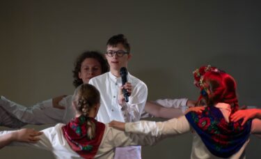 Młodzież w białych koszulach i czerwonych spodniach lub spódnicach tworzy krąg, trzymając się za ramiona. W tle chłopak śpiewa do mikrofonu.