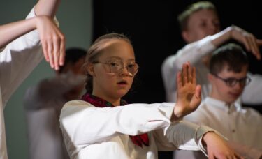 Grupa młodzieży z Integracyjnej Grupy Teatralnej wykonuje układ taneczny do spektaklu "Spacerkiem po Krakowie". Stoją tyłem w kręgu, kładąc sobie dłonie na ramionach. Widoczne są tradycyjne, kolorowe chusty i wianek, które są elementami ich ludowych strojów.
