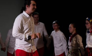 Uśmiechnięty chłopak wykonuje scenę. W tle młodzież obtacza go w kółki